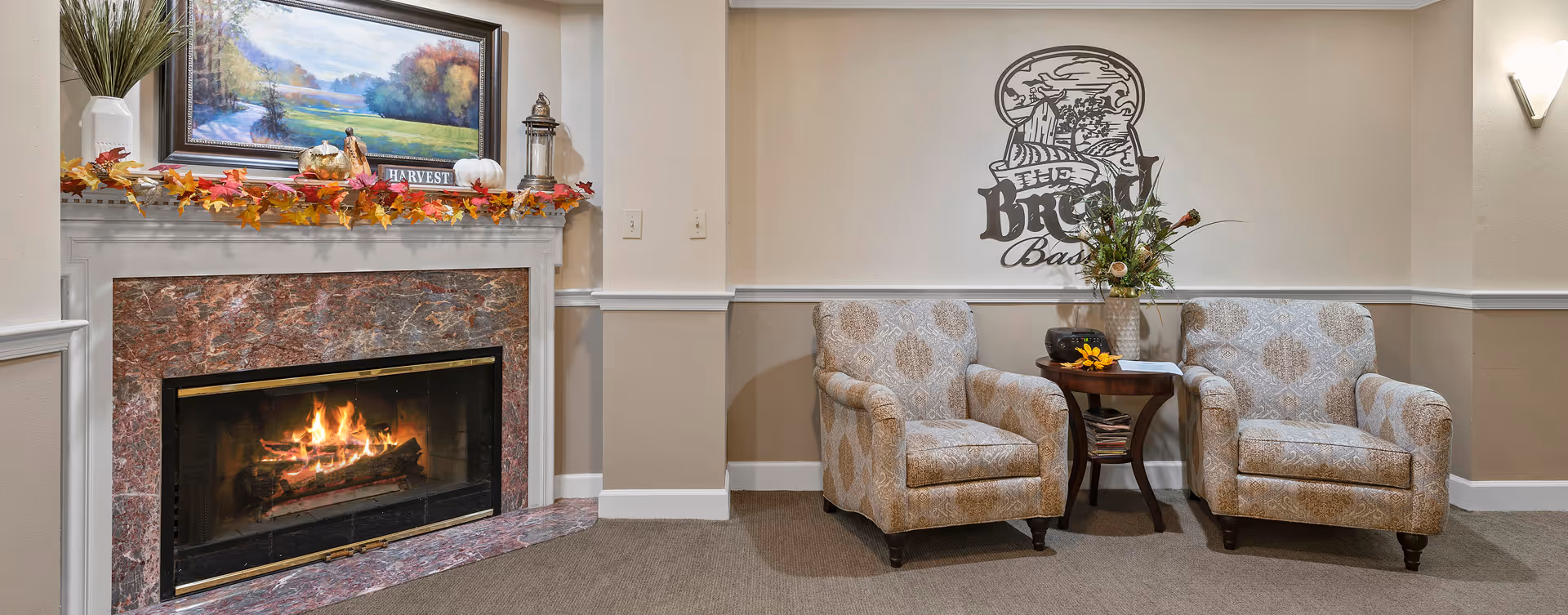 A cozy seating area with two patterned armchairs and a small wooden side table with a vase of flowers and a radio. To the left, there is a lit fireplace with a marble surround and a white mantel decorated with autumn leaves, pumpkins, a lantern, and a framed landscape painting. The wall behind the chairs has a decorative sign that reads 'The Bickford Basement'.
