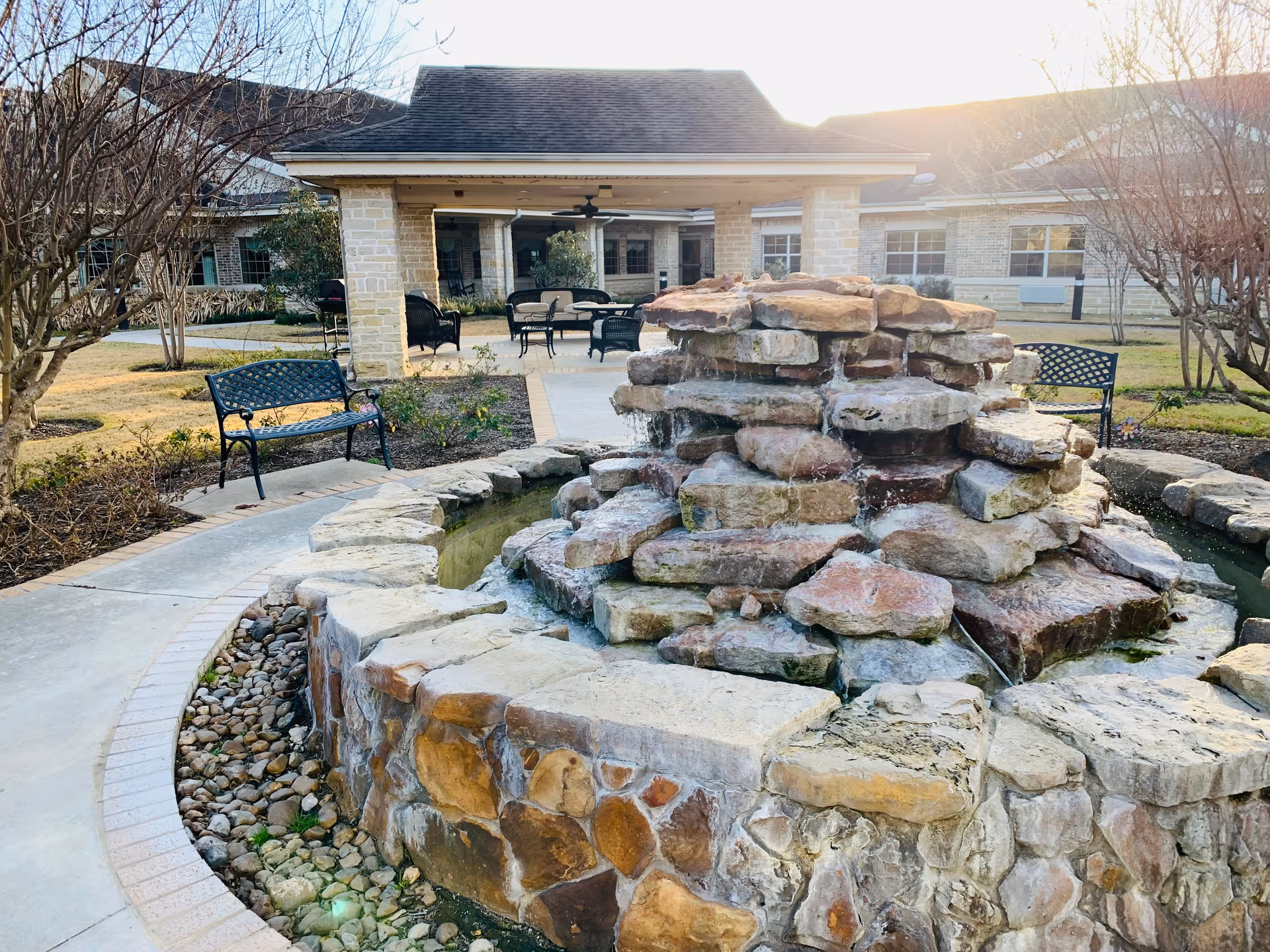 Stone tiered fountain in a landscaped outdoor courtyard with benches and a covered patio in the background.
