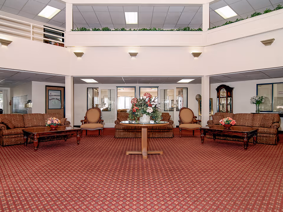 Spacious seating area in a senior living facility with patterned red carpet, two brown sofas, two beige armchairs, two wooden coffee tables with flower arrangements, a round table with a large floral centerpiece in the middle, and a grandfather clock against the wall. The room has a high ceiling with a balcony railing and overhead fluorescent lighting.