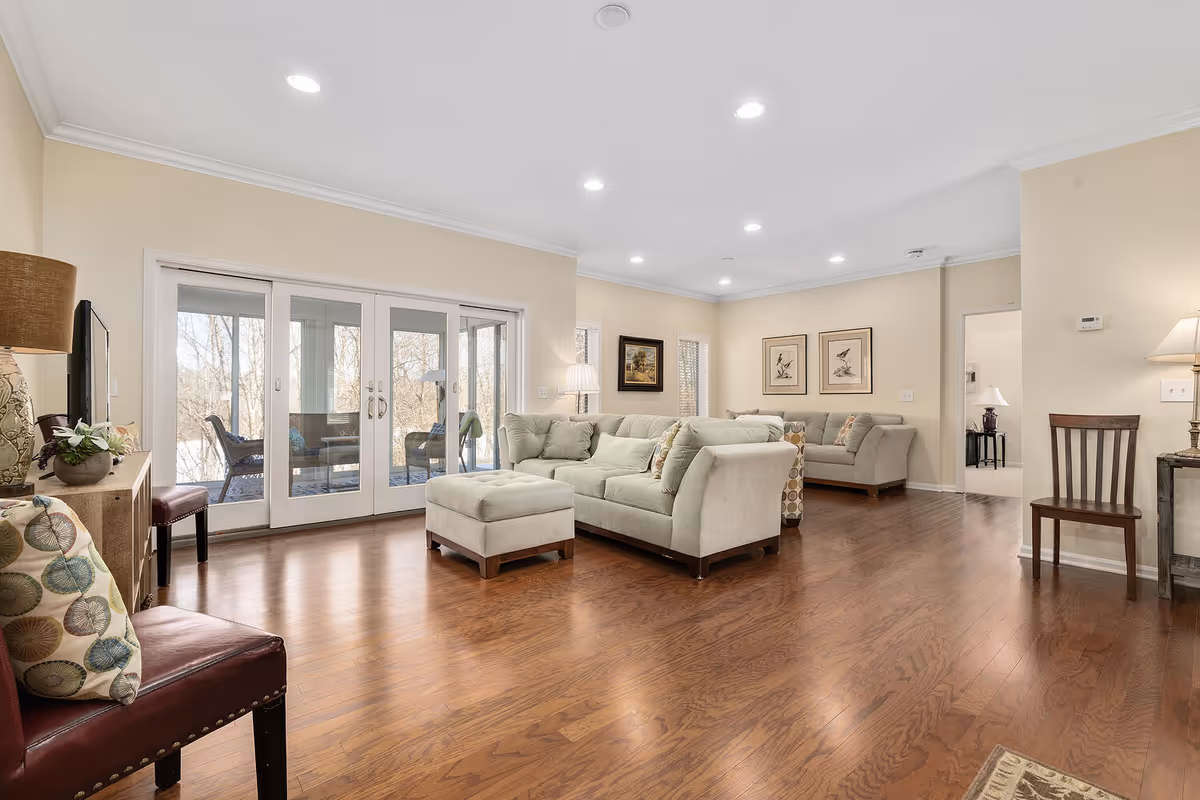 A spacious living room with light beige walls and wooden flooring. The room features a large light gray sectional sofa with matching ottoman, a smaller matching sofa, and several chairs with patterned cushions. There are framed artworks on the walls and a floor lamp beside the sectional. Large glass doors lead to an enclosed porch area with outdoor seating, and natural light fills the room.