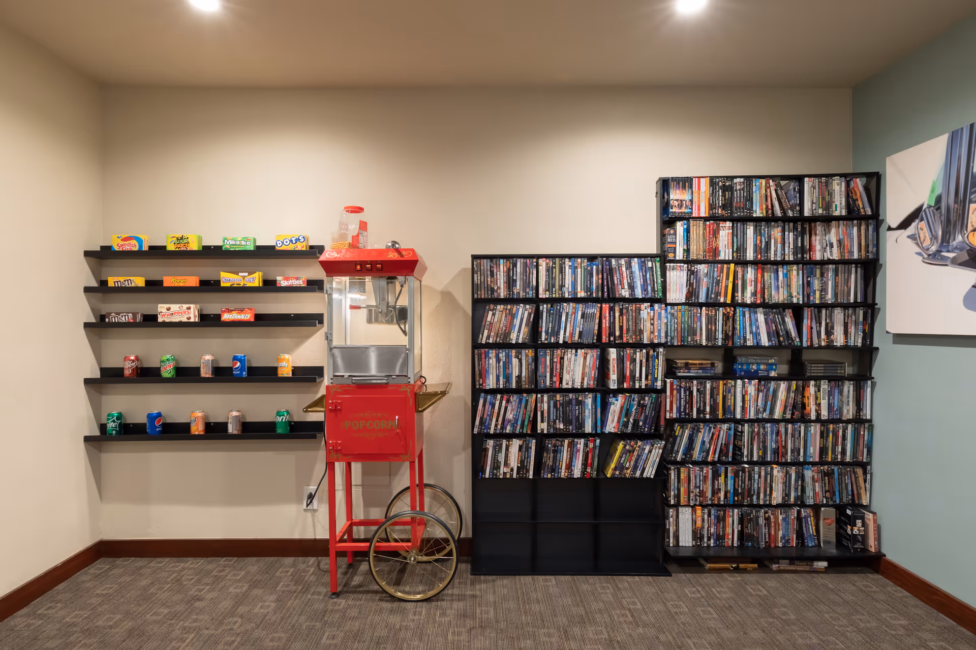 A cozy entertainment corner featuring a red vintage-style popcorn machine, shelves stocked with various candy boxes and soda cans, and two large black racks filled with DVDs against a beige wall with carpeted floor.