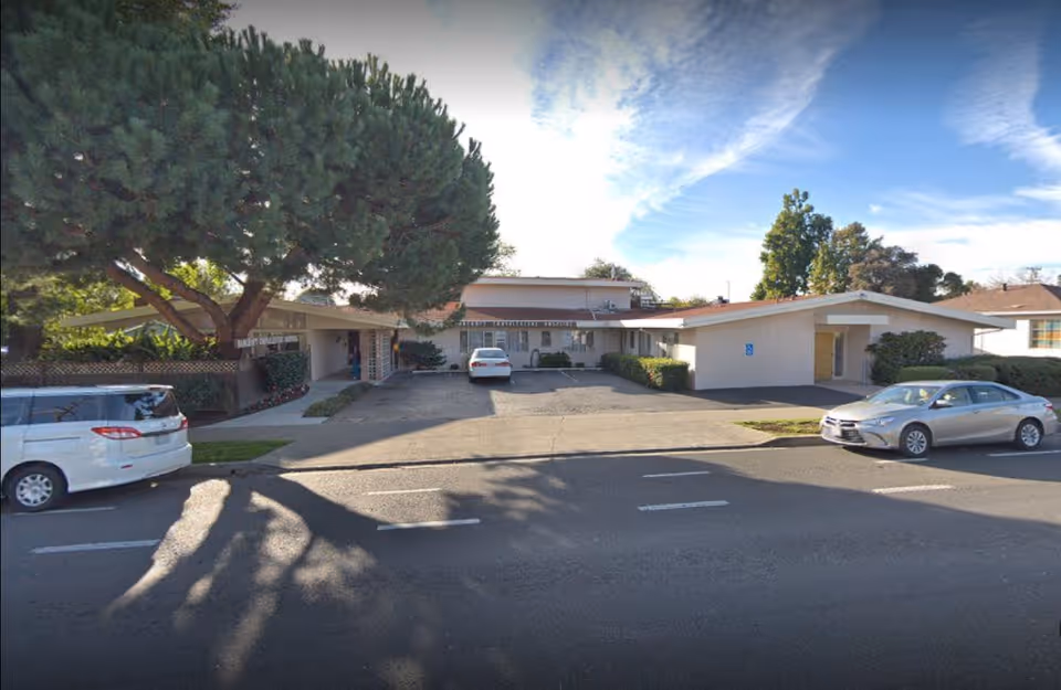 Street view of a one-story healthcare building with a parking area, parked cars, and large trees in front.