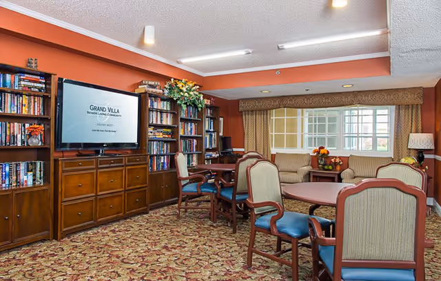 A cozy senior living community room with wooden bookshelves filled with books, a flat-screen TV mounted on the wall displaying 'Grand Villa Senior Living Community', several tables with chairs, and a seating area with armchairs near a large window with curtains. The room has warm orange walls and patterned carpet.
