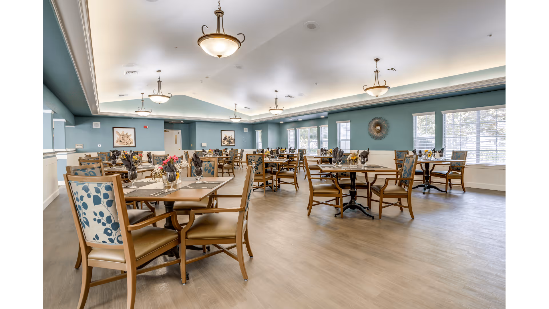 A spacious dining room with multiple wooden tables and chairs arranged neatly. Each table is set with placemats, glassware, and napkins, with small floral centerpieces. The room features light blue walls, large windows allowing natural light, and ceiling lights with decorative fixtures.