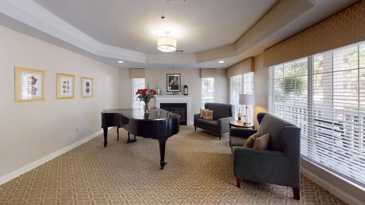 A bright and spacious living room in a senior living facility featuring a black grand piano with a vase of red flowers on top, two gray upholstered armchairs with decorative pillows, a small round side table with a lamp, large windows with blinds allowing natural light, a fireplace with decorative items on the mantel, and framed artwork on the walls.