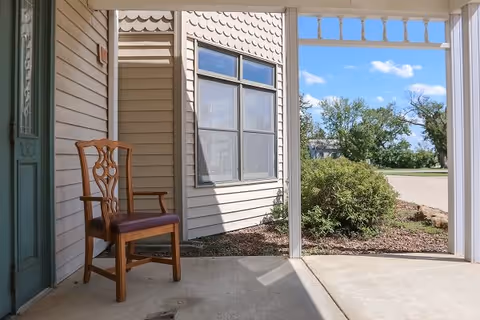 A wooden chair with a cushioned seat placed on a covered porch area outside a building. The porch has beige siding and a window, with a view of bushes, trees, and a clear blue sky in the background.
