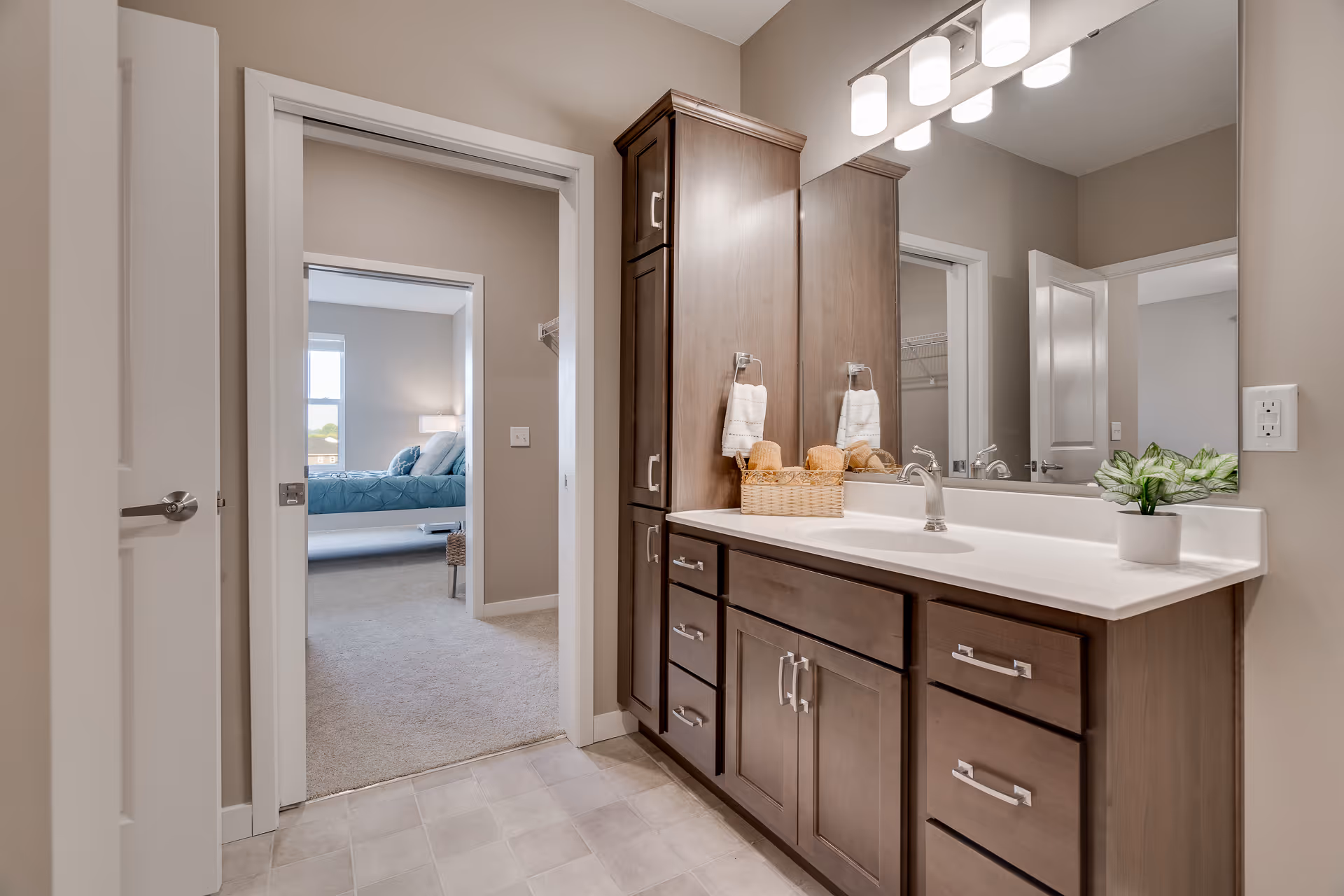 A bathroom vanity area with a large mirror, wooden cabinets, and a white countertop with a sink. There are two hand towels hanging on the wall, a basket with rolled towels, and a small potted plant on the counter. The bathroom opens into a bedroom with a bed covered in a blue quilt and a window letting in natural light.