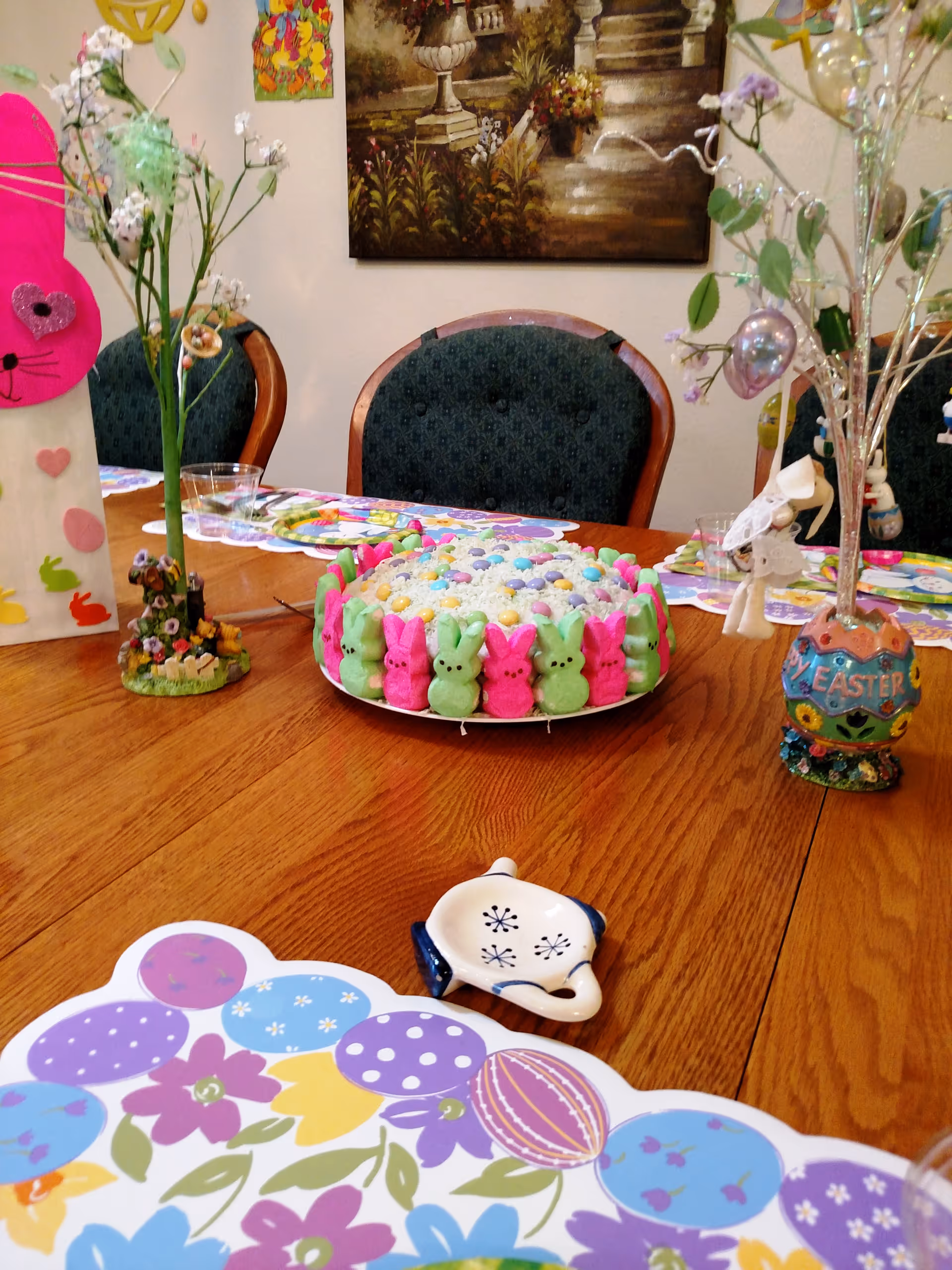 A wooden dining table decorated for Easter with a colorful cake surrounded by pink and green marshmallow bunny candies, floral placemats, and various Easter-themed decorations including a pink bunny figure and a decorative egg vase with branches.