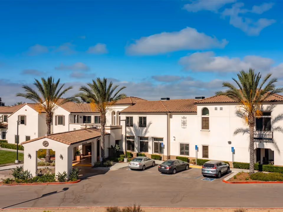 Exterior view of Westmont at San Miguel Ranch facility showing a two-story building with white walls and a red-tiled roof. There are several palm trees in front of the building and a covered entrance area. Three cars are parked in the parking lot, including two in handicapped spaces. The sky is blue with some clouds.
