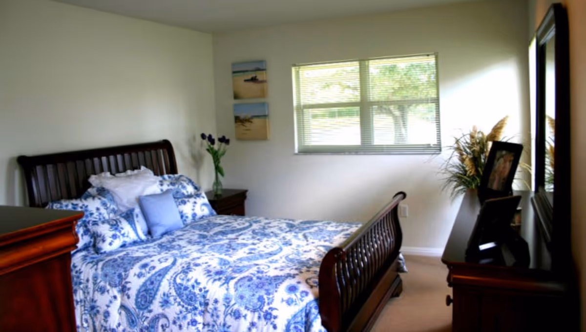 A bedroom with a wooden bed frame and a bed covered in a blue and white floral patterned comforter and pillows. There is a wooden dresser with a mirror on the right side, decorated with framed photos and a plant. A window with blinds lets in natural light, and two small beach-themed paintings hang on the wall next to the window.