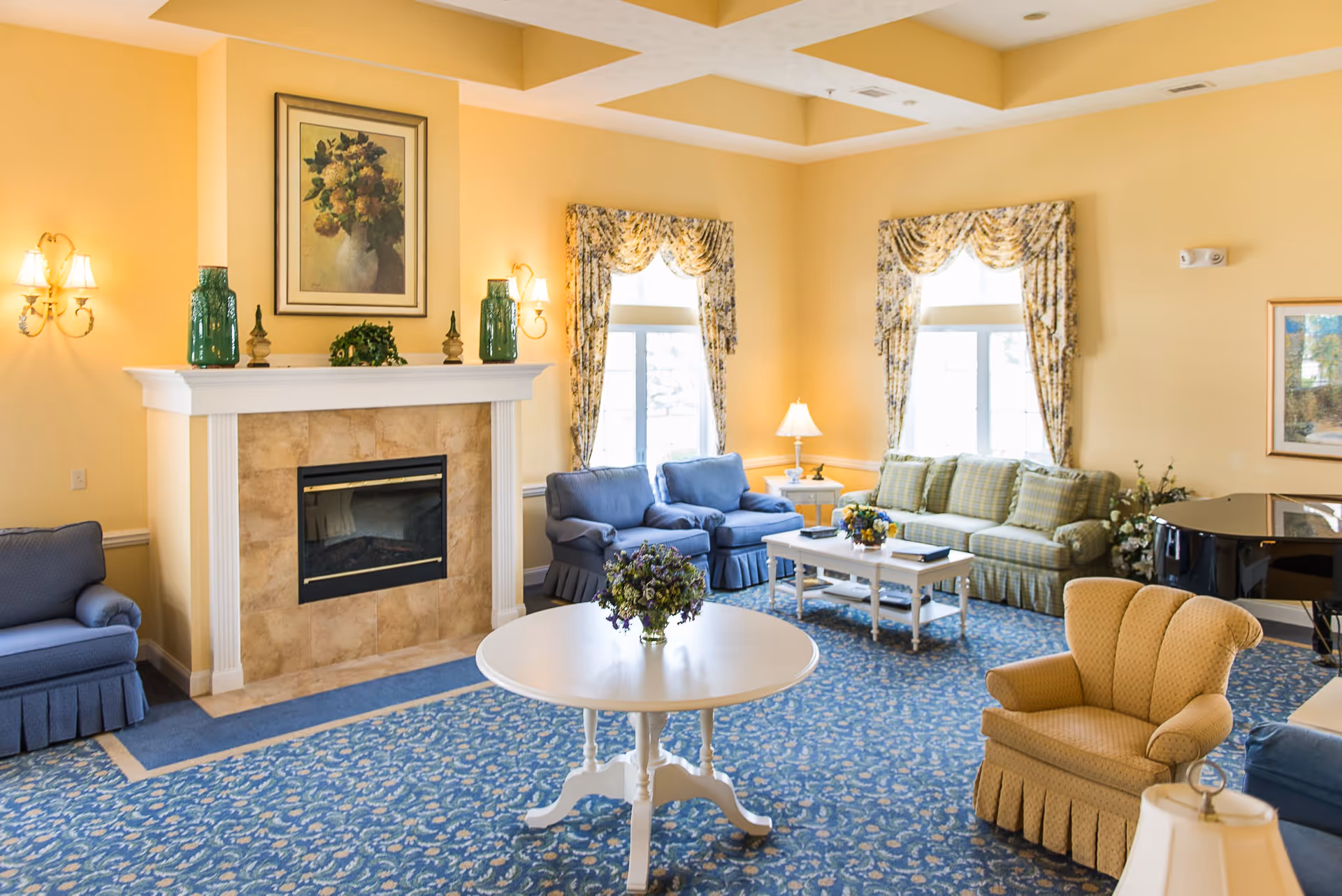 A cozy lobby area featuring a fireplace, comfortable seating with blue and yellow tones, decorative curtains, and a round table with a floral arrangement.