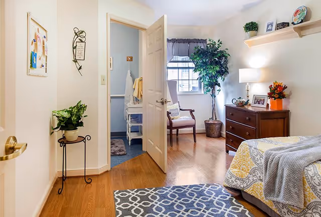A cozy bedroom in a senior living facility with a bed covered in a yellow and gray patterned quilt and a gray blanket. A wooden dresser with a lamp, framed photos, and a flower arrangement sits against the wall. A window with a valance lets in natural light, next to a wooden chair and a tall potted plant. The door to an adjacent bathroom is open, showing a sink with towels. A small plant on a stand and wall decorations are also visible.