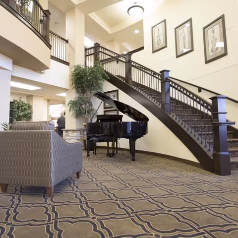 Interior view of a senior living facility featuring a grand staircase with dark wooden railings, a black grand piano, patterned carpet, a cushioned armchair, potted plants, and framed artwork on the wall.