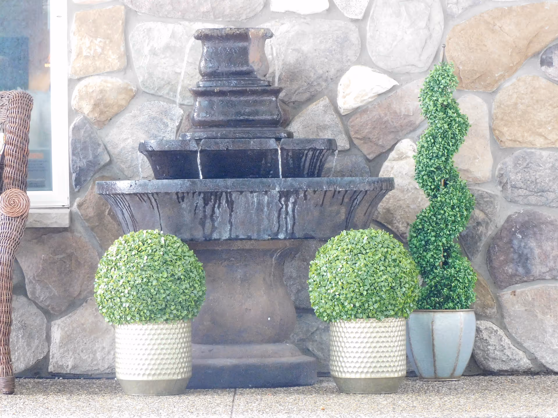 A decorative water fountain made of dark stone with water flowing down multiple tiers, placed against a stone wall. In front of the fountain are two round green potted plants in textured white pots and one tall spiral-shaped green plant in a blue pot. Part of a wicker chair is visible on the left side.