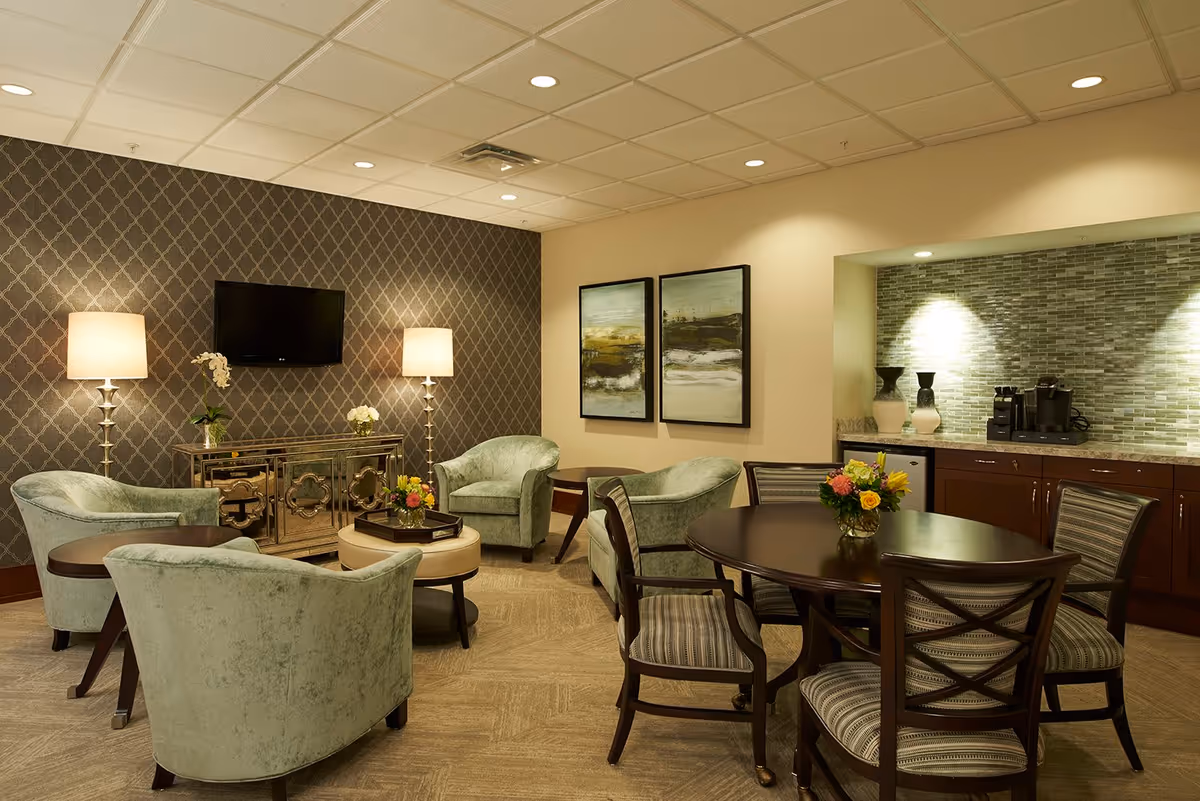 A cozy living room area with four light green upholstered armchairs arranged around a small round ottoman and a dark wood side table. Two tall floor lamps flank a wall-mounted flat-screen TV above a mirrored cabinet. On the right side, there is a round dining table with four striped cushioned chairs and a flower arrangement on top. The back wall features two framed abstract paintings and a small kitchenette area with a coffee maker, vases, and a tiled backsplash.