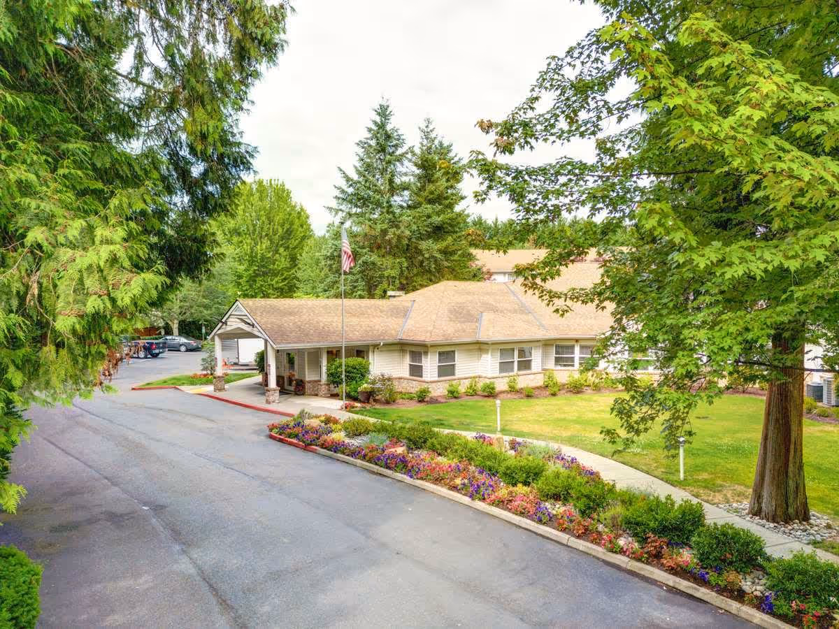 Exterior view of a single-story building with a covered entrance, surrounded by green trees and a well-maintained garden with colorful flowers along the driveway.