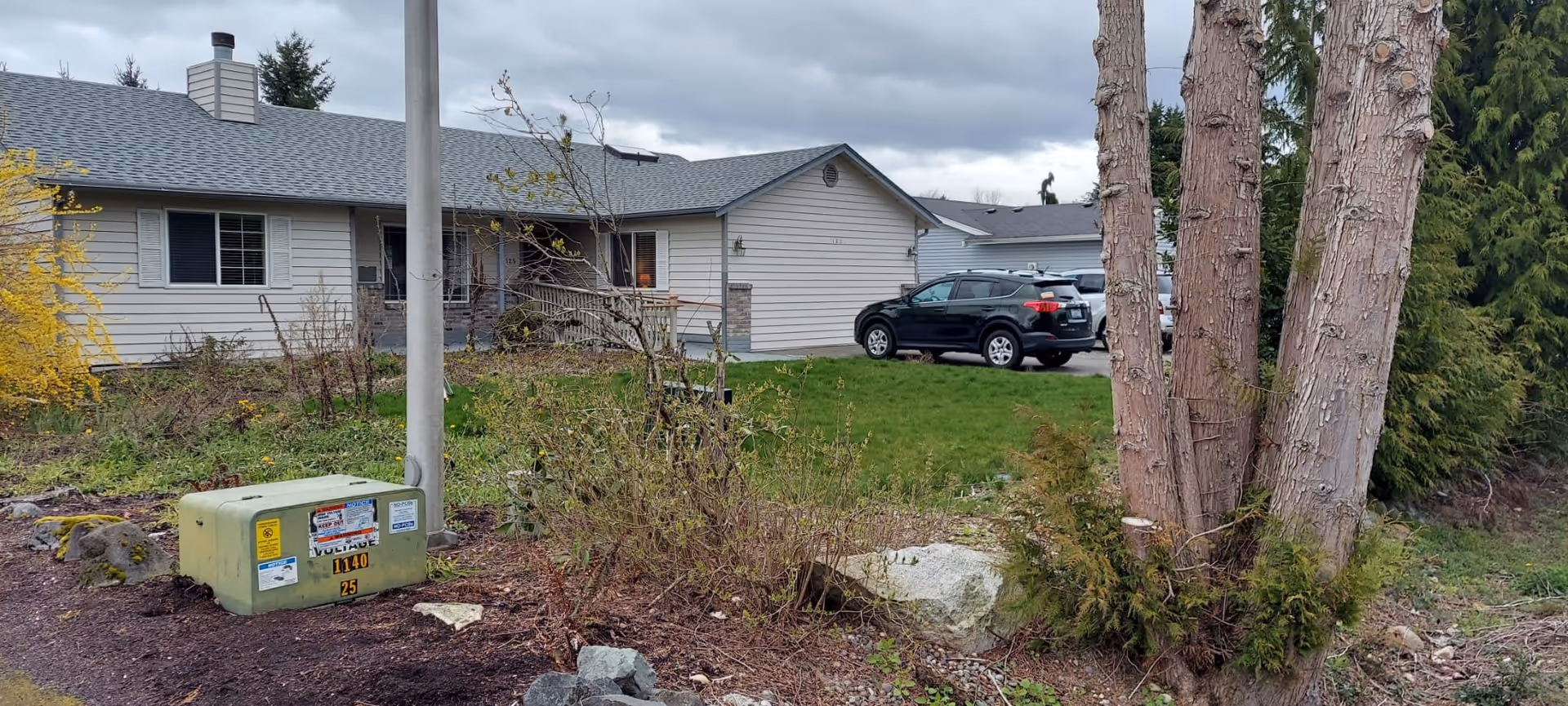 Single-story residential building with a driveway and parked SUV, lawn, trees and a utility box in the foreground under an overcast sky.