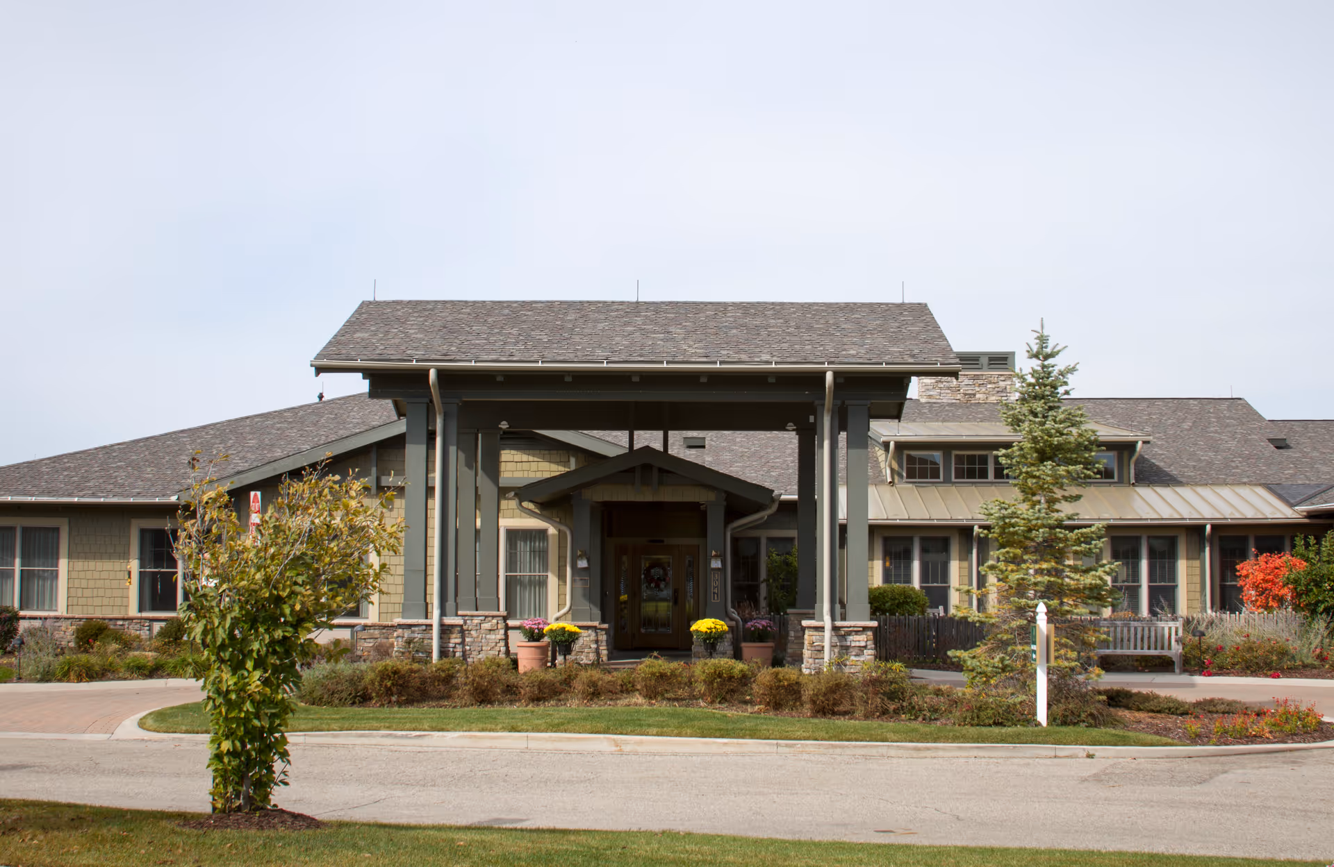 Front exterior view of a single-story building with a covered entrance supported by columns. The building has a shingled roof, stone accents, and several windows. There are potted plants with yellow flowers near the entrance and landscaped greenery including bushes and small trees in front.