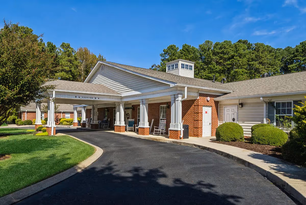 Exterior view of Kennesaw Village senior living facility showing a covered entrance with white columns and rocking chairs, surrounded by well-maintained landscaping and trees under a clear blue sky.