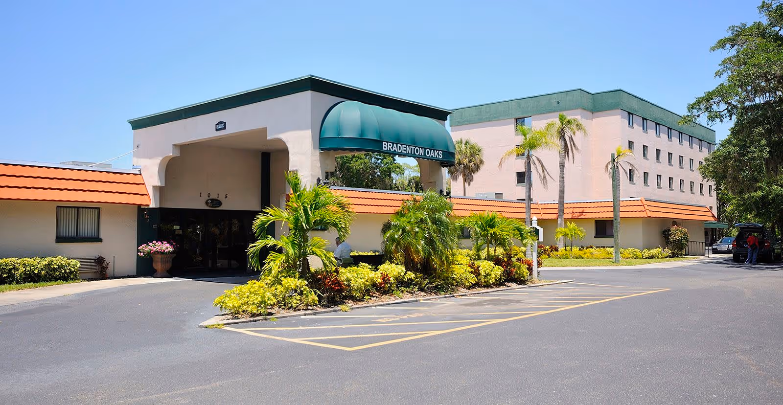 Front entrance of a senior living facility with a green awning reading 'Bradenton Oaks', surrounding landscaping and parking lot.