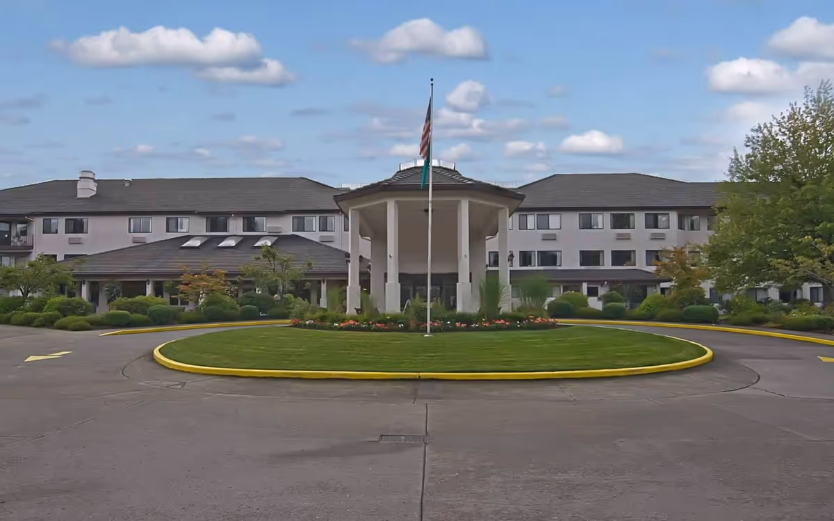 Front entrance of a multi-story senior living building with a circular driveway, landscaped lawn and a flagpole.