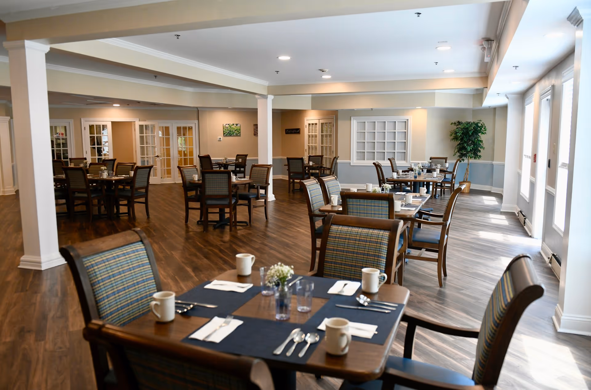A spacious dining room with multiple wooden tables and chairs arranged neatly. Each table is set with placemats, cups, utensils, and small flower vases. The room has wooden flooring, light-colored walls, and large windows letting in natural light. There are decorative plants and framed pictures on the walls.