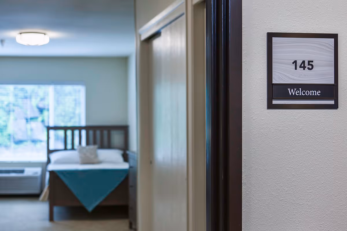 View down a hallway into a bedroom with a bed, a pillow, and a teal blanket. A window with blinds and an air conditioning unit are visible in the background. On the wall near the hallway entrance is a sign with the number 145 and the word 'Welcome'.