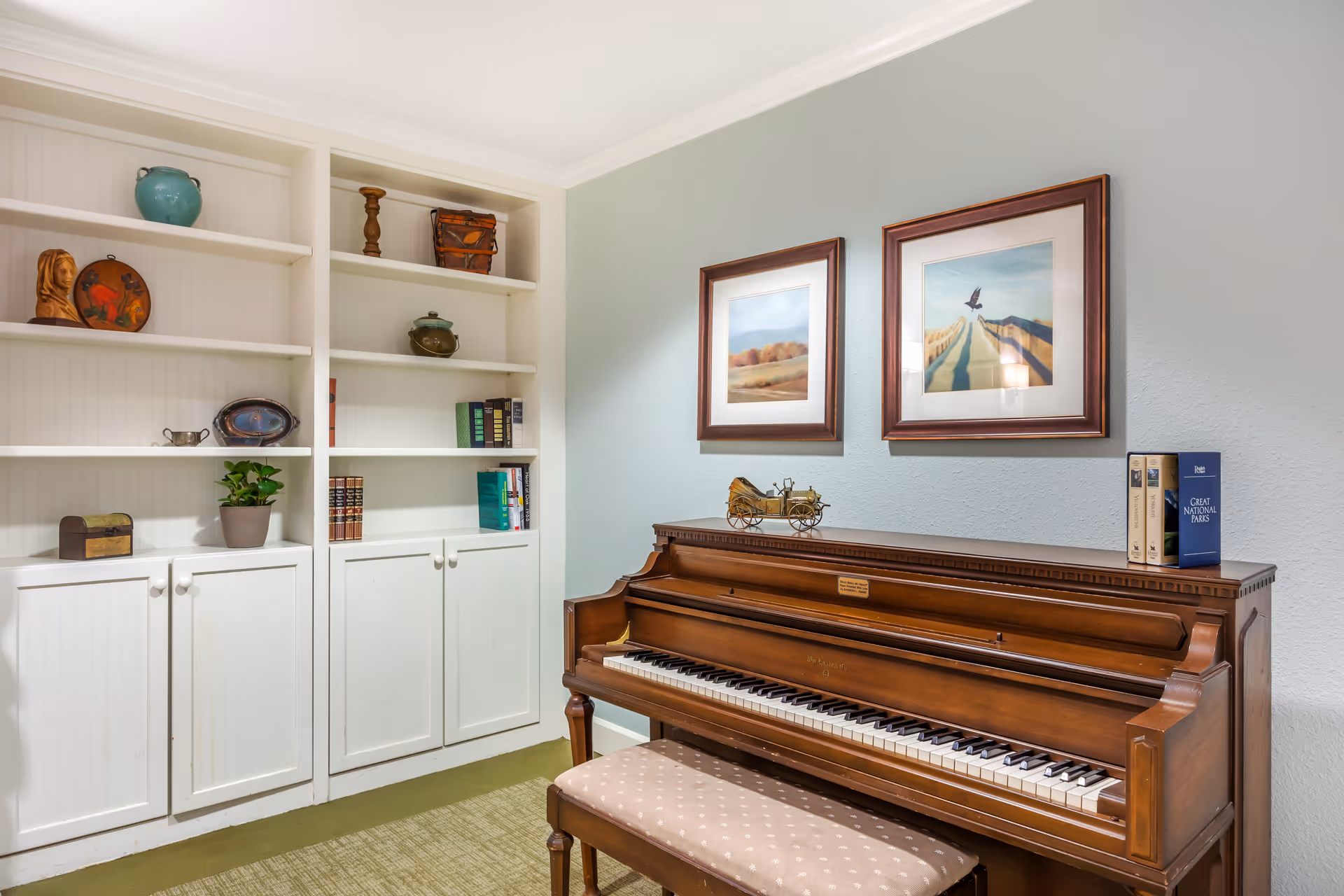 A cozy interior room featuring a wooden upright piano with a cushioned bench in front. Above the piano are two framed landscape paintings. To the left, there is a white built-in bookshelf with various decorative items, books, and a small potted plant. The walls are painted light blue and the floor is carpeted in green.