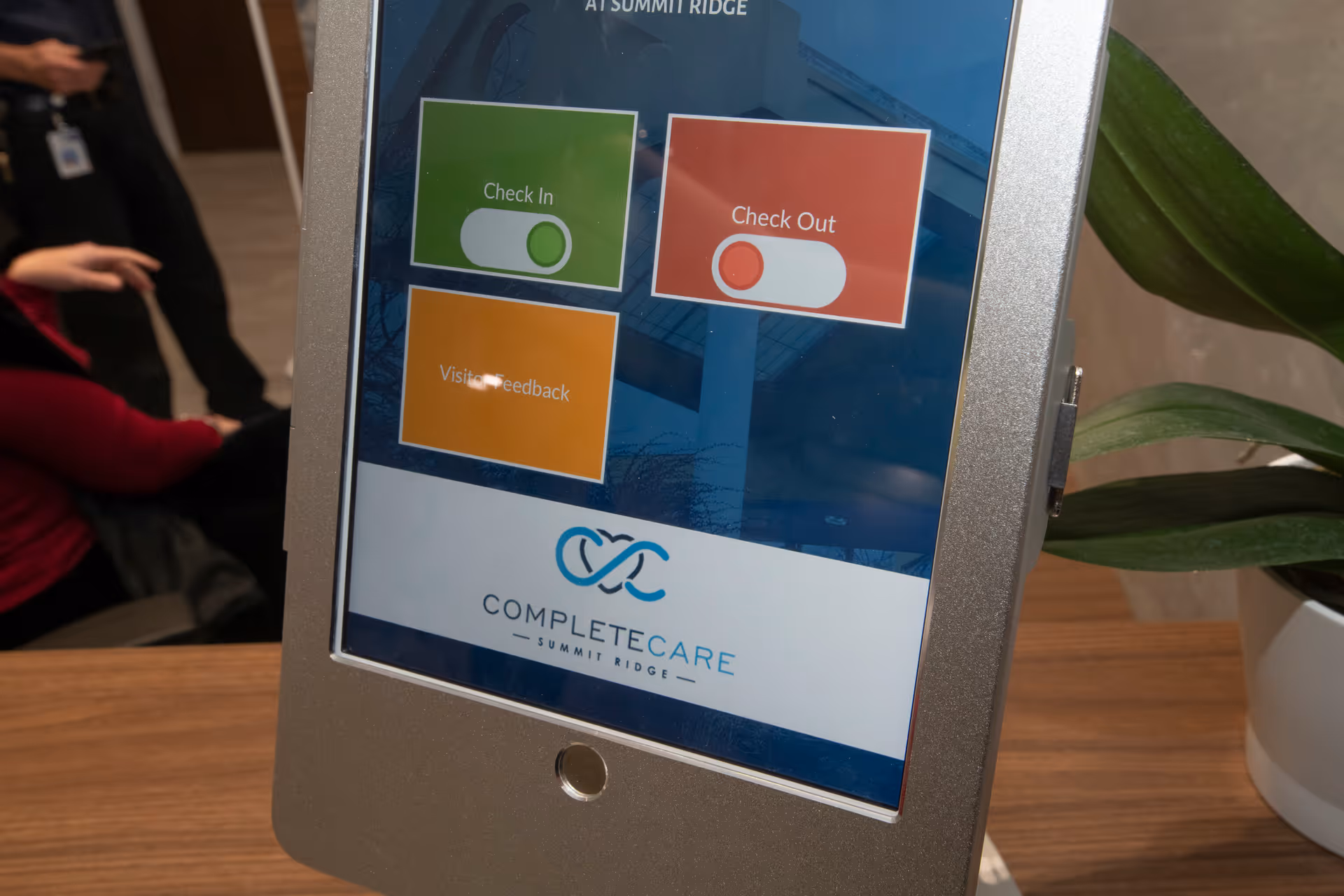 A digital check-in kiosk screen at Complete Care at Summit Ridge displaying options for Check In, Check Out, and Visitor Feedback. The kiosk is placed on a wooden surface next to a green plant in a white pot. In the background, a person in a red shirt and another person in black clothing are partially visible.