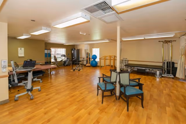 A spacious rehabilitation room with wooden flooring, exercise equipment including stationary bikes, therapy balls, parallel bars, and treatment tables. There are also a few chairs arranged near a support column, and the room is well-lit with ceiling lights and windows.