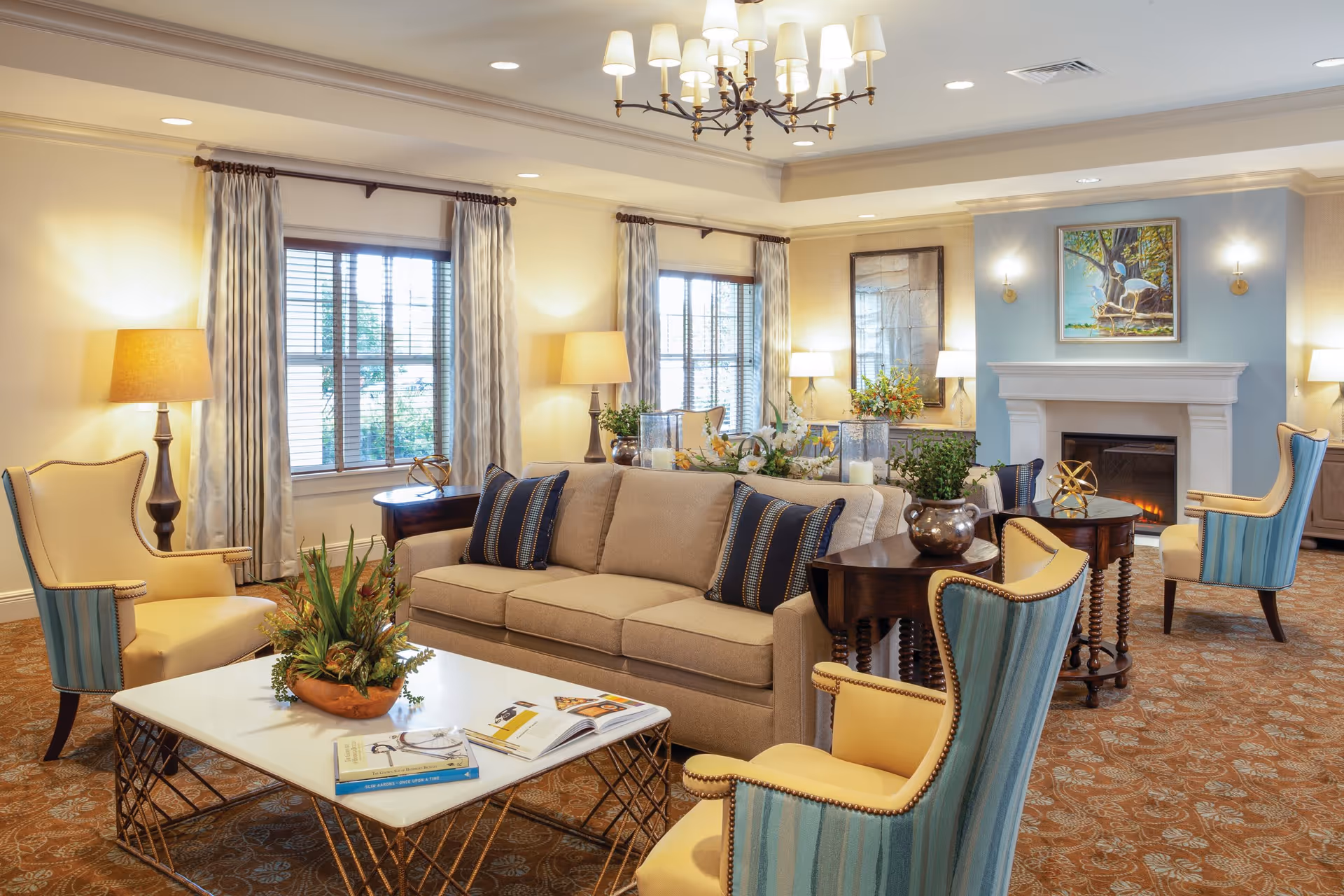 A cozy and elegant living room with a beige sofa adorned with dark blue pillows, four yellow and blue striped armchairs, a white coffee table with books and a plant centerpiece, two side tables with plants, warm lighting from lamps and a chandelier, large windows with curtains, and a fireplace with a painting above it.