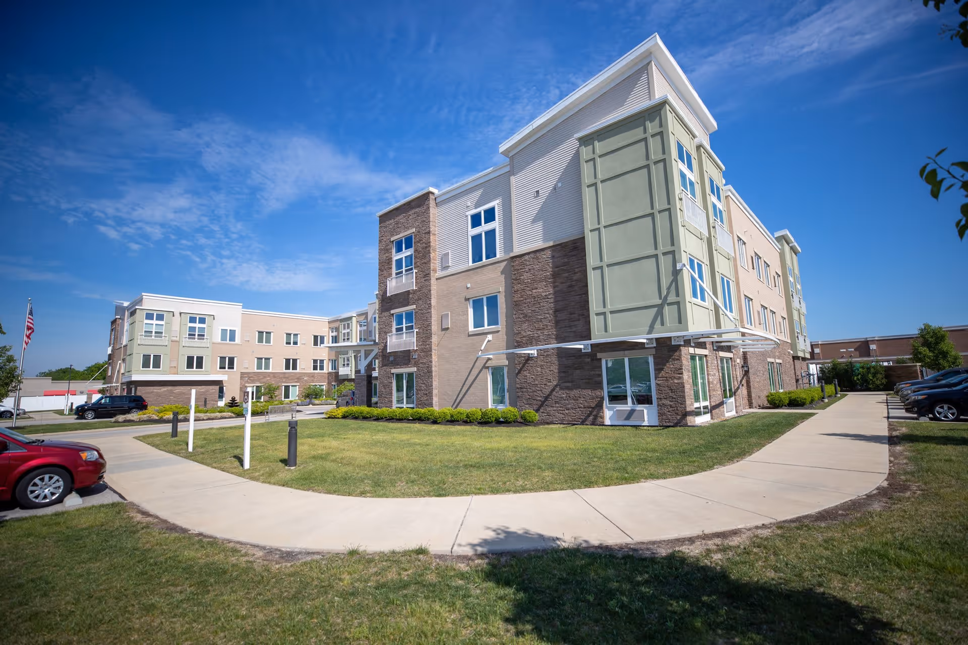 Exterior view of a modern three-story senior living facility building with a mix of beige, green, and stone facade under a blue sky. There is a curved sidewalk in front, a well-maintained lawn, several parked cars, and an American flag on a flagpole.