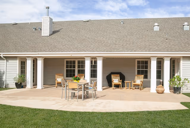 Covered back patio of a senior living building with a dining table, wooden chairs, potted plants, and a lawn.