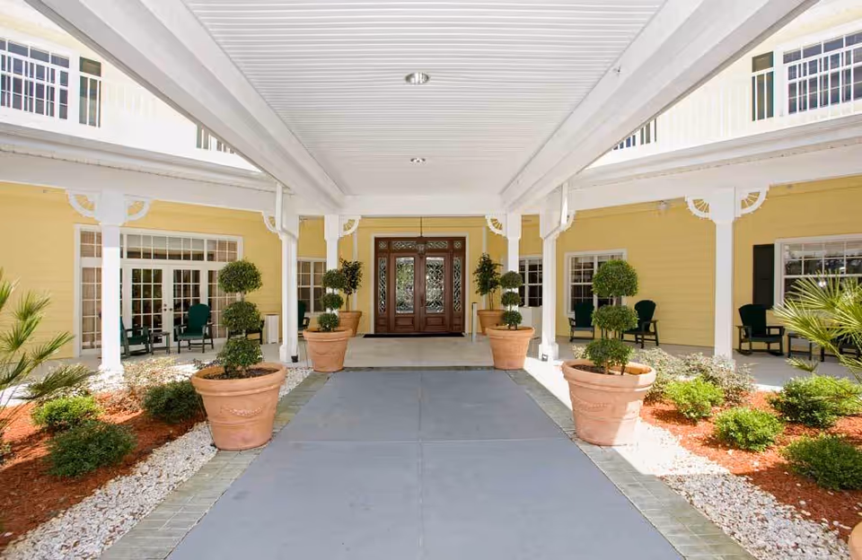 Covered entrance area of a senior living facility with large double wooden doors, yellow exterior walls, potted topiary plants, and green chairs on the porch.
