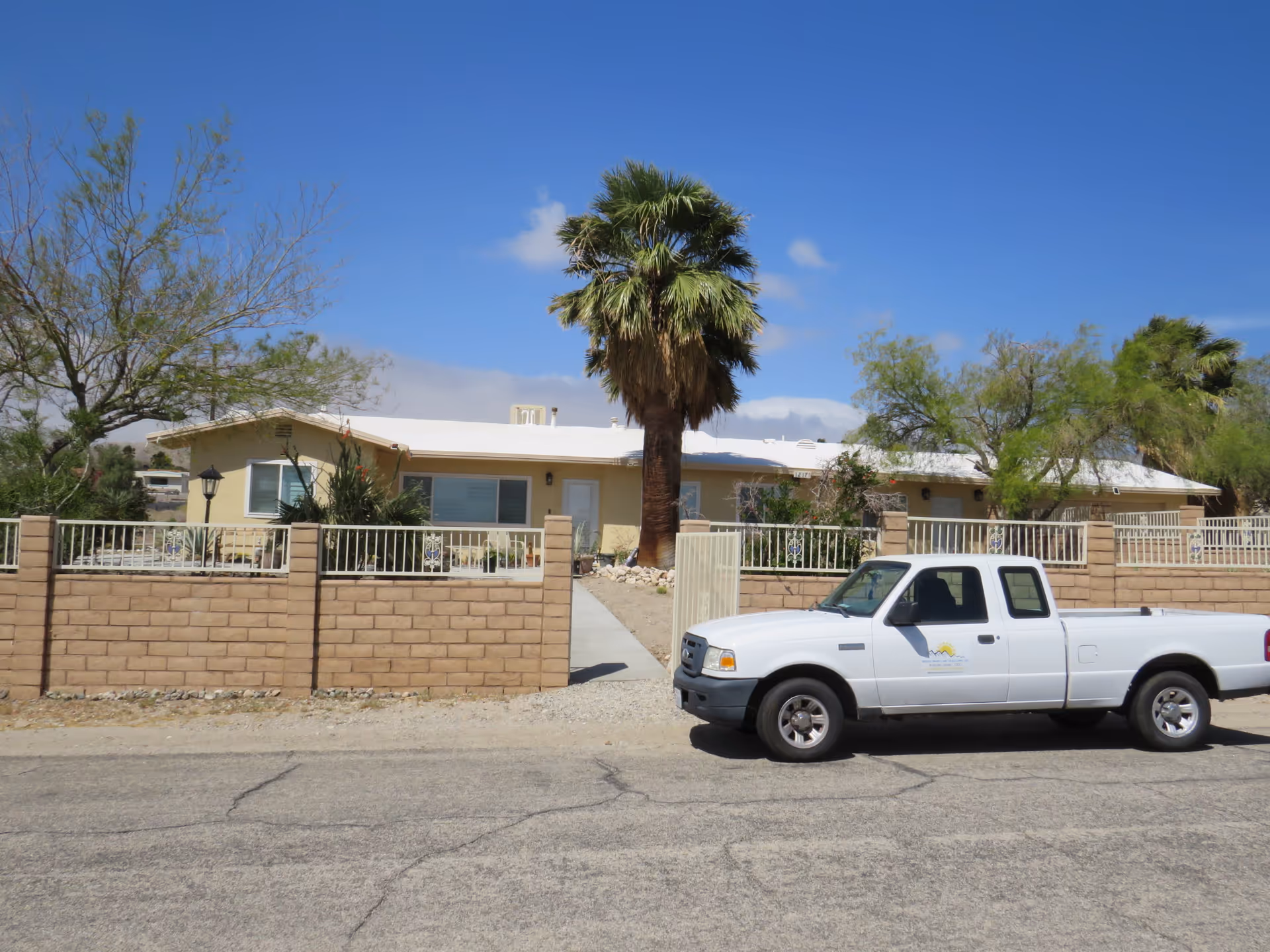 Exterior view of a single-story building with a beige brick fence and a white metal gate. A tall palm tree is in front of the building, and a white pickup truck is parked on the street in front. The sky is clear and blue with a few clouds.