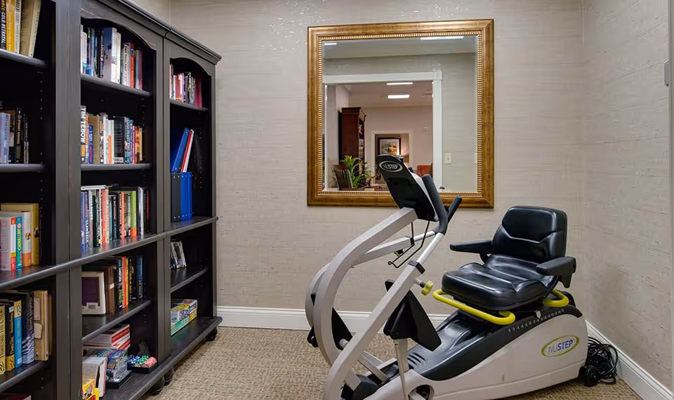 A small room with a recumbent exercise bike positioned near a large wall mirror. To the left, there is a tall dark wooden bookshelf filled with books and some board games. The room has beige textured walls and carpeted flooring.