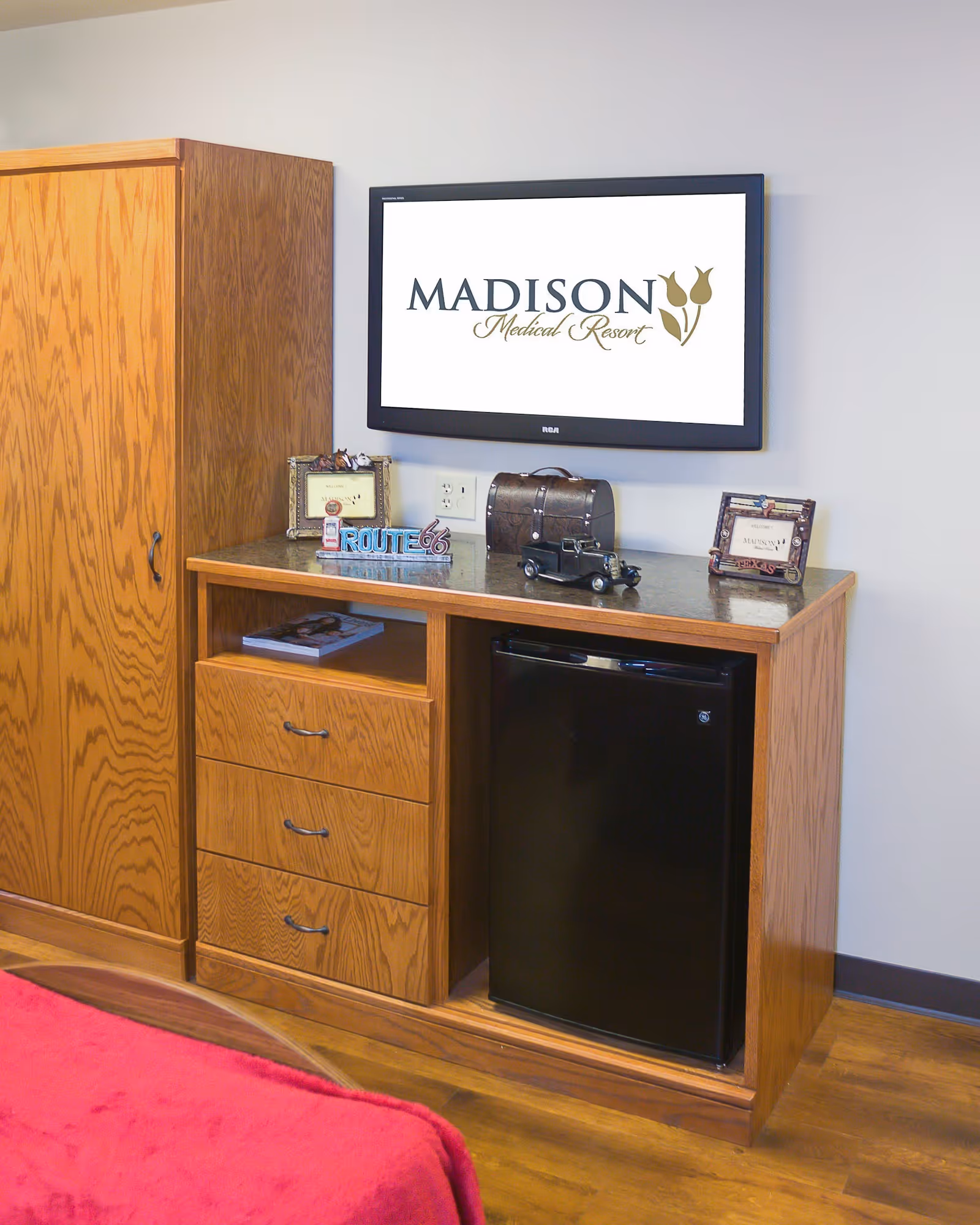 Wall-mounted TV displaying 'Madison Medical Resort' above a wooden dresser with drawers, a mini fridge and decorative items, with the corner of a red bed visible.