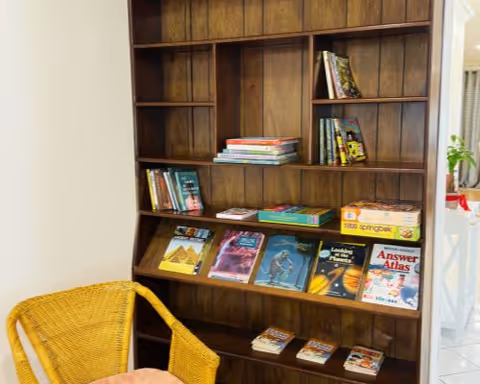 Wooden bookcase stocked with books and board games next to a wicker chair in a light room.