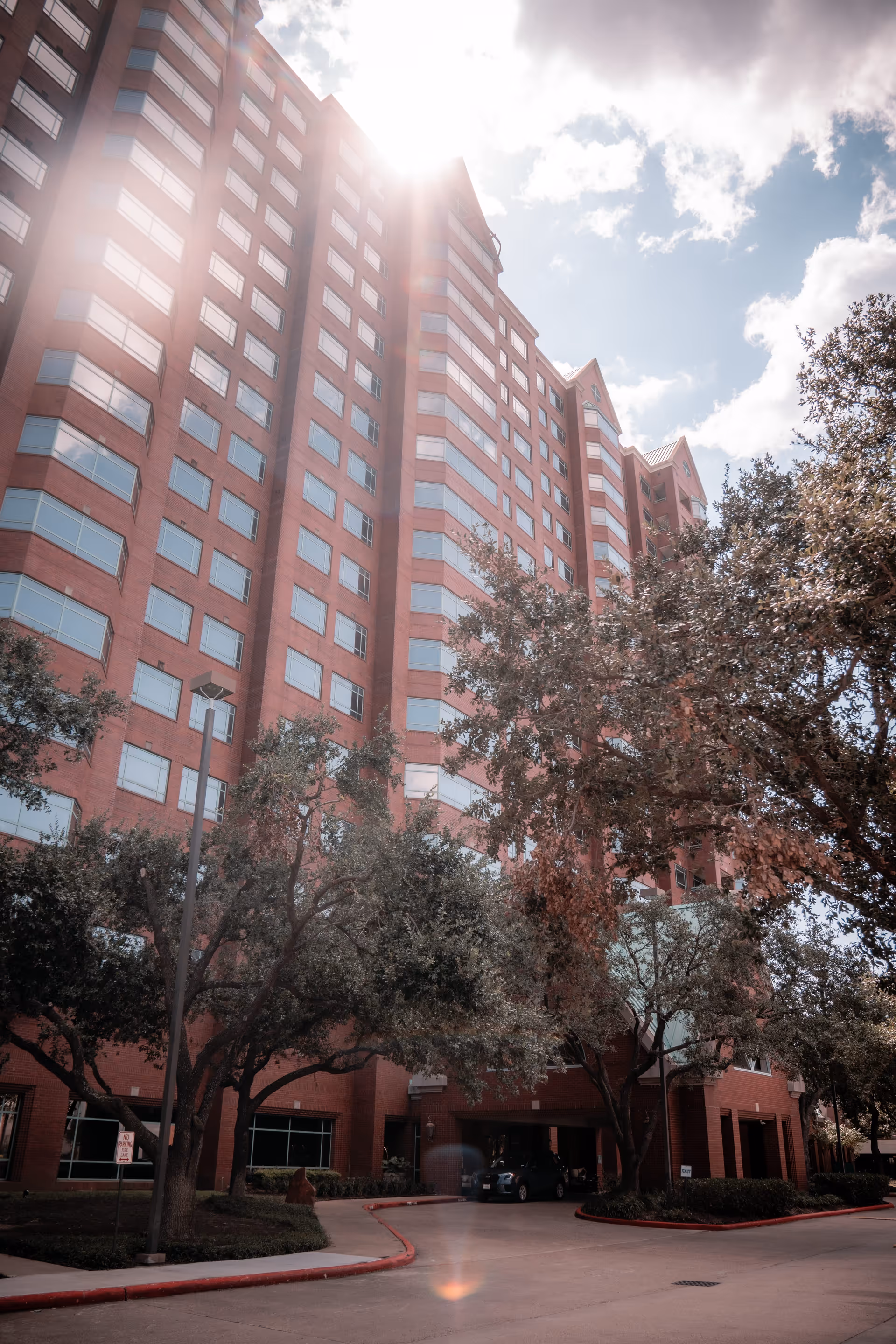 Red-brick high-rise building entrance with trees, a driveway, and a bright sunlit sky.