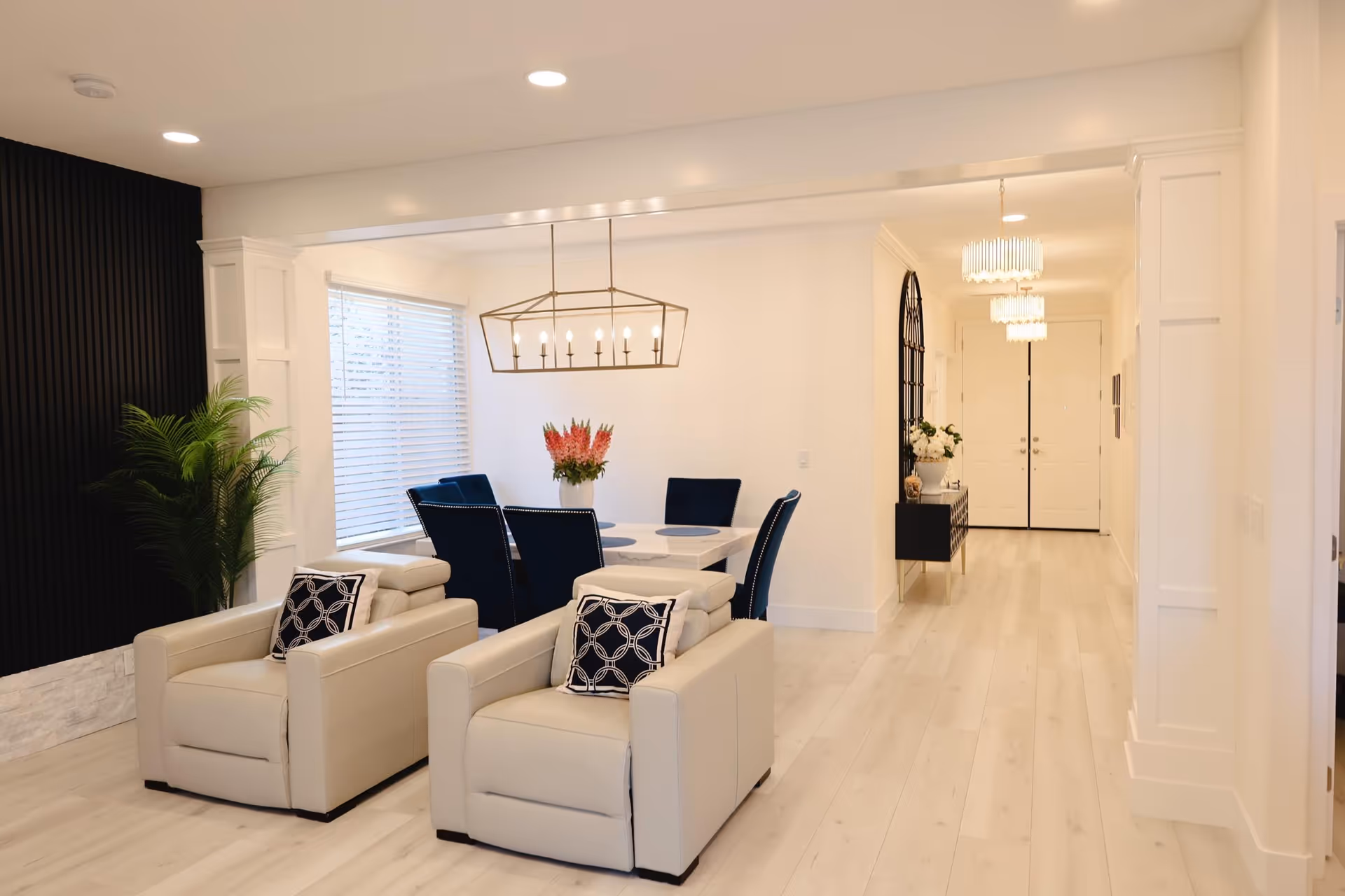 A bright and modern interior space featuring two beige armchairs with patterned cushions in the foreground, a dining area with a white table and six dark blue chairs, a large window with blinds, a geometric chandelier above the table, a green potted plant against a black accent wall, and a hallway with white double doors and pendant lights in the background.