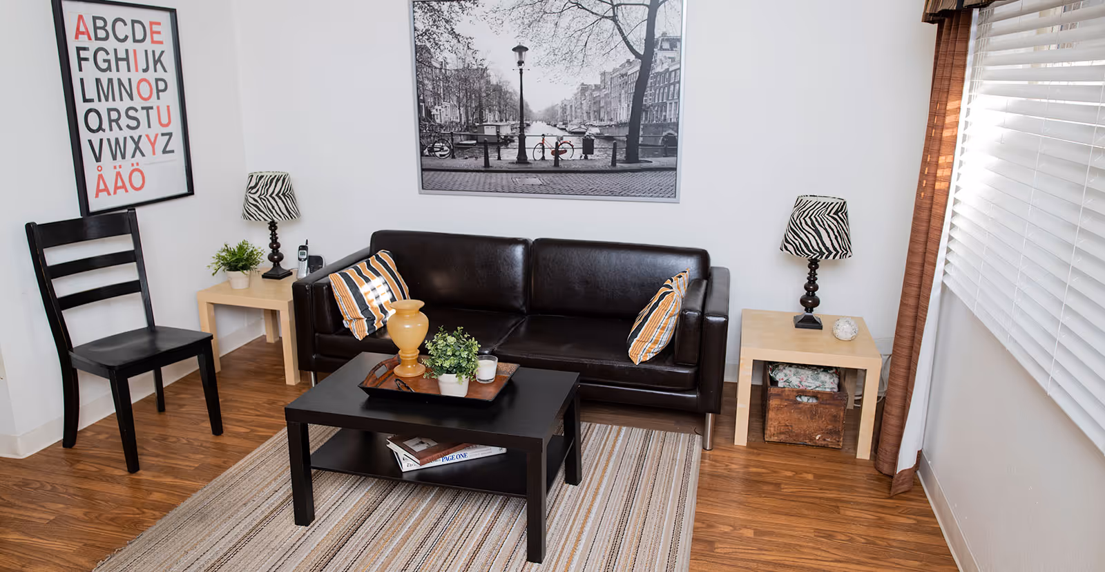 A cozy living room with a black leather sofa adorned with two striped pillows, a black coffee table with decorative items and books underneath, two light-colored side tables each with a zebra-patterned lamp, a black wooden chair, a framed alphabet poster on the wall, and a large black and white photograph of a canal scene. The room has wooden flooring and a window with white blinds and brown curtains.