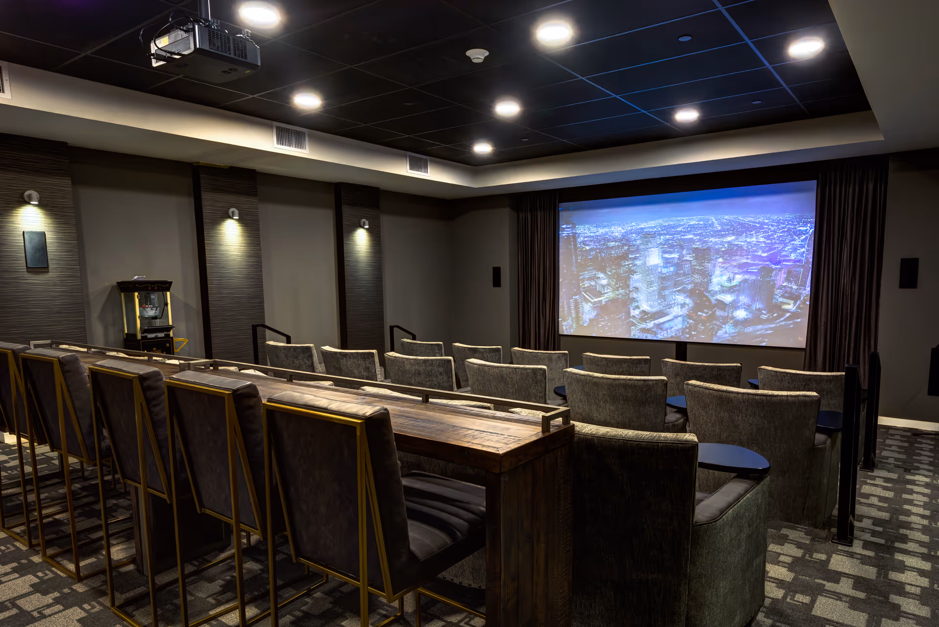 A modern theater room with rows of comfortable seating facing a large screen displaying a cityscape at night. The room has dim lighting, a popcorn machine in the corner, and a ceiling-mounted projector.