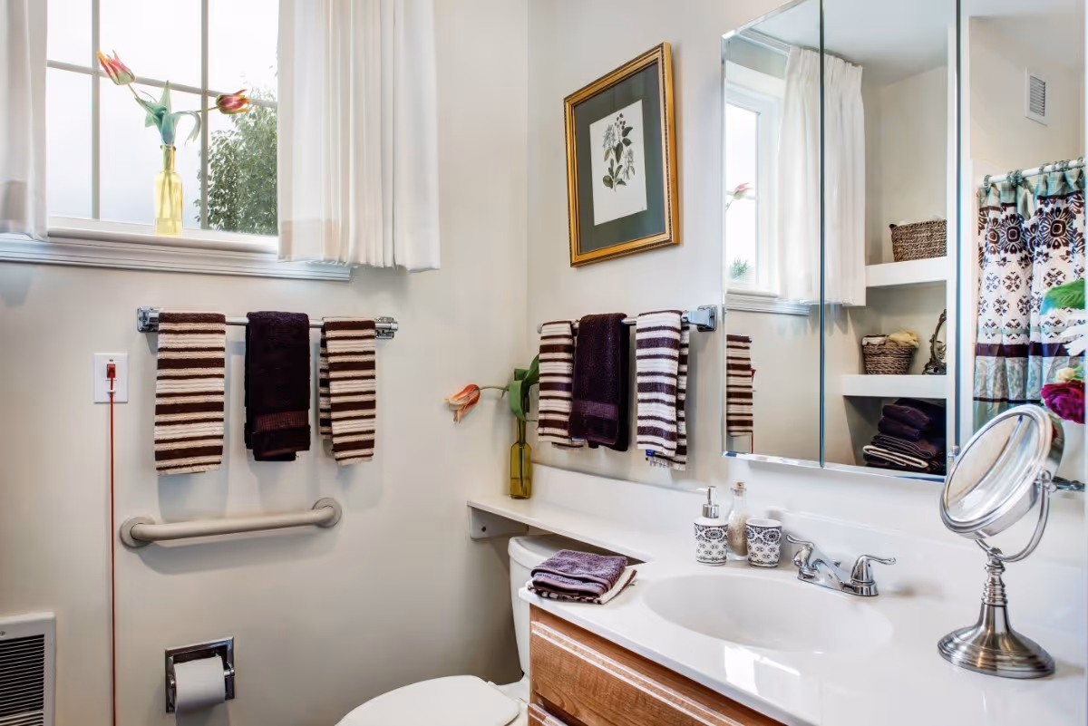 Bright bathroom with a sink and mirror, toilet, striped towels on racks, and decorative accents like a framed print and flowers.