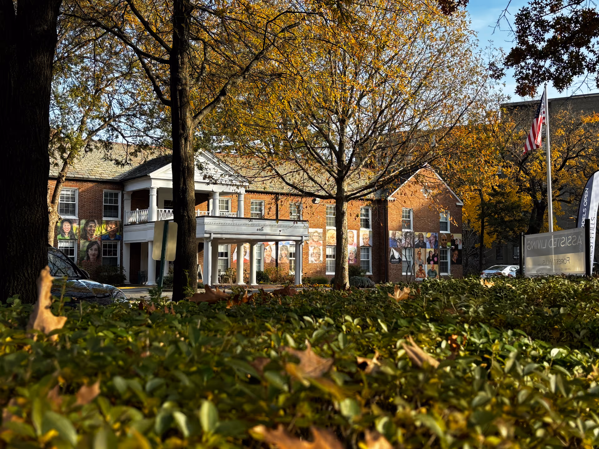 Exterior view of a brick building with white columns and a covered entrance, surrounded by trees with autumn foliage and green bushes in the foreground. The building has large portraits of various people displayed on its walls. An American flag is visible on a flagpole near a sign that reads 'ASSISTED LIVING FOREST HILLS'.