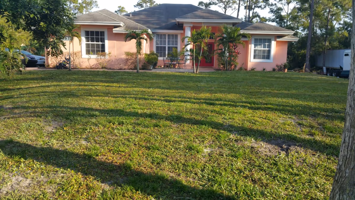 Front exterior view of a single-story pink house with a gray roof, red front door, and several windows. The house is surrounded by a large grassy lawn with some small palm trees and other plants near the entrance. Trees and a parked vehicle are visible in the background.