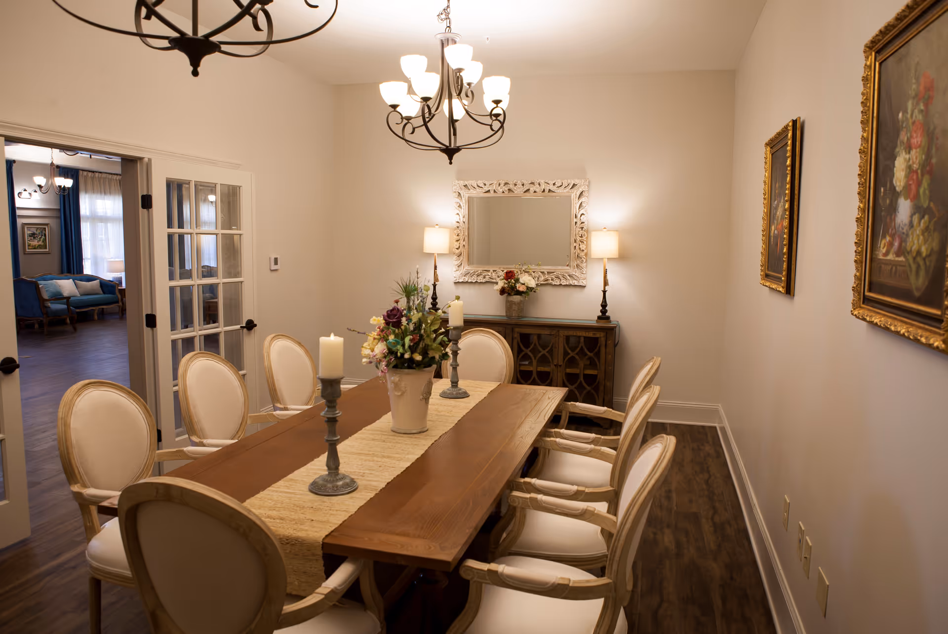 A dining room with a wooden table surrounded by eight upholstered chairs. The table is decorated with a beige runner, two candle holders with white candles, and a floral arrangement in a vase. On the far wall, there is a decorative mirror flanked by two table lamps on a wooden sideboard. The room has hardwood flooring, beige walls, and two framed paintings on the right wall. A doorway with glass-paneled double doors leads to another room with blue curtains and a blue sofa.