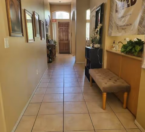 A long hallway with tiled floor leading to a wooden door with a window above it. The hallway walls are decorated with framed pictures on the left and a cushioned bench with plants and decorative items on the right. Natural light is coming through windows on the right side.