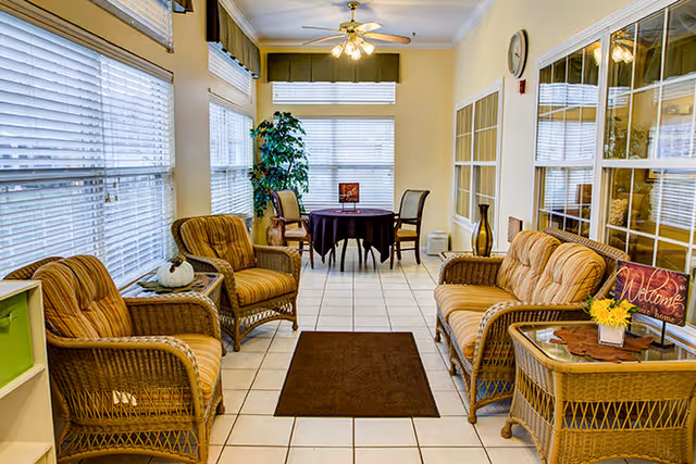A bright sunroom with large windows covered by white blinds. The room features wicker furniture with striped cushions, including two armchairs, a loveseat, and a glass-top side table. A small brown rug is placed on the tiled floor. At the far end, there is a round table covered with a dark tablecloth and surrounded by four chairs. A potted plant is positioned near the table. A decorative sign that says 'Welcome' and a small vase with yellow flowers are on the side table.