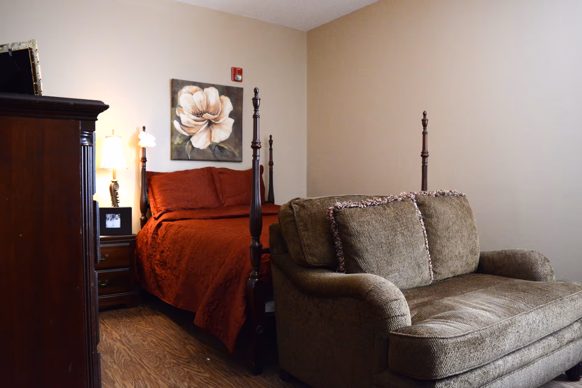 Room with a four-poster bed covered in red bedding, a bedside lamp and nightstand with wall art, and a green loveseat in the foreground.