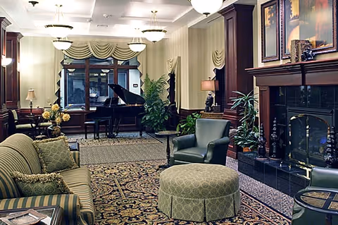 A cozy living room area with a patterned carpet, green upholstered chairs, a round ottoman, a striped sofa, and a grand piano near the window. The room features a fireplace with framed artwork above it, several plants, and soft lighting from ceiling fixtures and lamps.